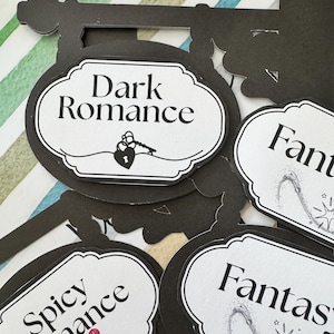 May include: Black and white decorative signs with the text "Dark Romance", "Spicy Romance", and "Fantasy". The signs have a black border and a white interior with a decorative shape. The "Dark Romance" sign has a lock and key illustration.