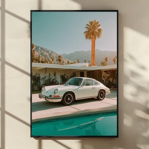 Könnte beinhalten: Gerahmter Druck mit einem weißen Oldtimer-Sportwagen vor einem Haus im Stil der Moderne. Die Szene umfasst einen Swimmingpool, Palmen und Berge unter einem klaren blauen Himmel. Das Auto hat schwarze Räder.