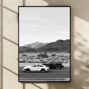 May include: Black and white framed print featuring two classic sports cars racing on a desert highway. The cars are a white and a black model, set against a backdrop of mountains and a clear sky. A stylish piece for car enthusiasts.