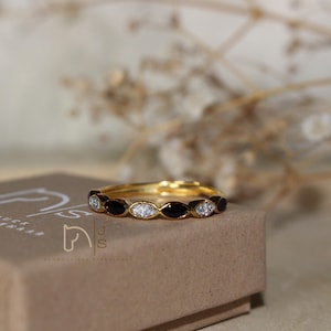 Pode incluir: Um anel de tom dourado com um padrão alternado de incrustações de esmalte preto em forma de marquise e pedras claras brilhantes. O anel está posicionado sobre uma caixa de cartão castanha com o logótipo branco JS e um ícone de cavalo.