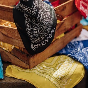 May include: A wooden crate overflowing with colorful bandanas. A black bandana with the word "forever" in white script is draped over the side. Other bandanas are blue, yellow, and red, with various paisley and decorative patterns.