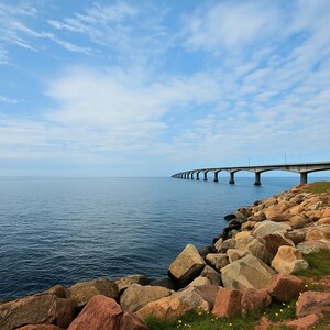Op de afbeelding: Een lange, grijze brug strekt zich uit over het blauwe water, met een rotsachtige kustlijn op de voorgrond. De lucht is helderblauw met witte wolken.
