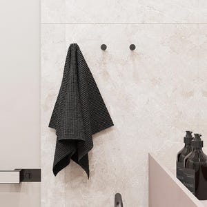 May include: A dark grey waffle-weave towel hangs from a black hook on a light-coloured tiled wall. Two black hooks are visible. The scene includes a glimpse of a sink and soap dispensers, suggesting a bathroom setting.