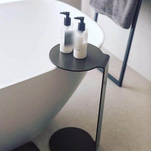 May include: A contemporary black metal side table with a circular top and base, designed for beside a bathtub. Two clear bottles with black pump dispensers are on the table, possibly for bath products. The table has a minimalist design.