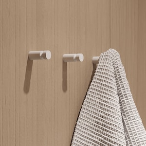 May include: Three white wall hooks with a cylindrical design are mounted on a light brown wooden surface. A white waffle-weave towel hangs from the third hook. The hooks are evenly spaced and create a minimalist aesthetic.