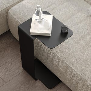 May include: A modern black side table with a unique C-shaped design. A clear glass bottle, a white book, and a small glass are on the tabletop. The table is positioned next to a light-colored sofa.