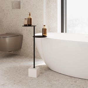 Può includere: Un tavolino laterale in metallo nero con due ripiani circolari, che contengono bottiglie marroni. Il tavolo ha una base quadrata bianca ed è posizionato accanto a una vasca da bagno bianca. Il bagno ha una combinazione di colori neutri.