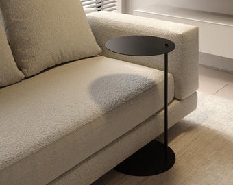 May include: A modern black side table with a round top and a slender stem. The table is positioned next to a beige textured sofa with matching pillows. The setting is a well-lit room with a neutral color palette, creating a minimalist aesthetic.