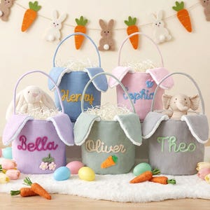 以下が含まれることがあります： パステルカラーのコーデュロイ製イースターバスケットのコレクション。それぞれにウサギの耳のアクセントとパーソナライズされた名前が付いています。バスケットには紙の草が詰められ、ぬいぐるみやニンジンのアップリケで飾られています。背景にはお祝いのイースターバナーが飾られています。