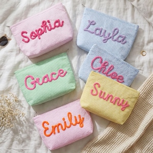May include: A collection of personalized cosmetic bags in various pastel colors, including pink, blue, green, and yellow. Each bag features a different name embroidered in a contrasting color. The bags are arranged on a white textured surface.