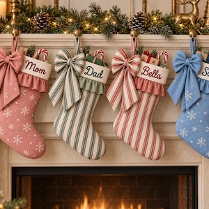 Op de afbeelding: Vier kerstsokken hangen op een rij, elk voorzien van een grote stoffen strik, een geplooide boord en gepersonaliseerd borduurwerk. De sokken tonen de namen Mom, Dad, Bella en Sophie, met patronen van sneeuwvlokken en verticale strepen in diverse kleuren.