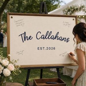 May include: A rectangular wedding guest book sign featuring a cream-coloured fabric surface in a gold frame. The text reads The Callahans and EST. 2026 in dark blue script and block letters. Handwritten notes are visible on the fabric surface.