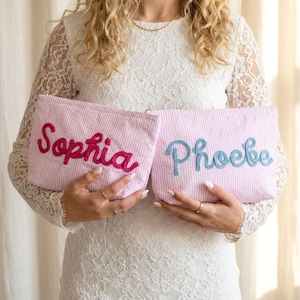 May include: Two pink seersucker pouches, each embroidered with a name. One pouch has "Sophia" in magenta, the other has "Phoebe" in blue. The pouches are rectangular and have a zipper closure.