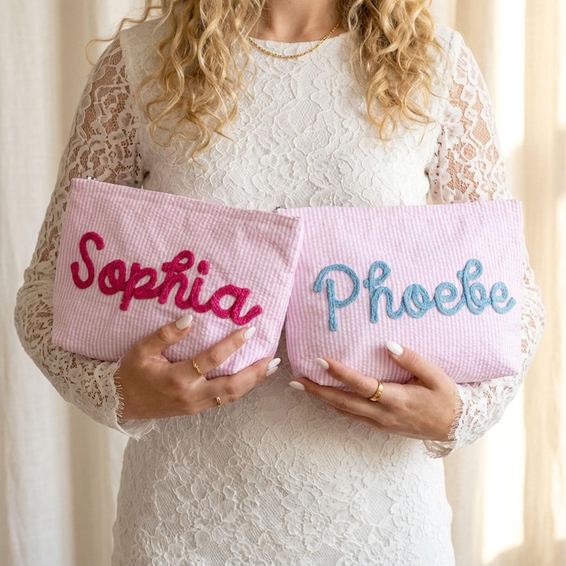 May include: Two pink seersucker pouches, each embroidered with a name. One pouch has "Sophia" in magenta, the other has "Phoebe" in blue. The pouches are rectangular and have a zipper closure.