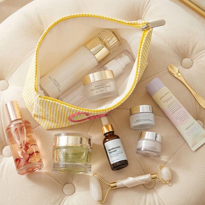 May include: A collection of skincare products, including bottles, jars, and tubes, arranged on a soft, cream-colored surface. A yellow and white striped cosmetic bag is open, revealing more products. A jade roller and a gold-colored spoon are also present.