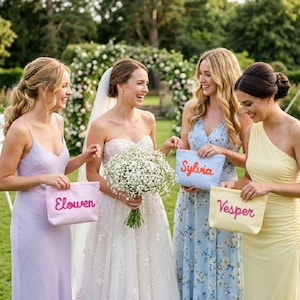 May include: Four women in pastel dresses holding personalized striped pouches. The bride wears a white gown and veil, holding a bouquet. The pouches are pink, blue, and yellow, each with a name embroidered in a contrasting color.