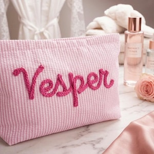 May include: Pink seersucker cosmetic bag with the name "Vesper" embroidered in a darker pink script. The bag is on a marble surface, with a rose, skincare products, and a white robe in the background.