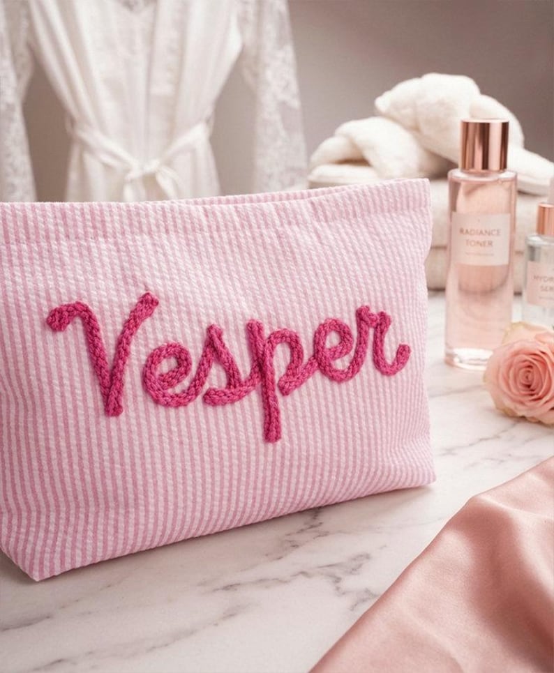 May include: Pink seersucker cosmetic bag with the name "Vesper" embroidered in a darker pink script. The bag is on a marble surface, with a rose, skincare products, and a white robe in the background.