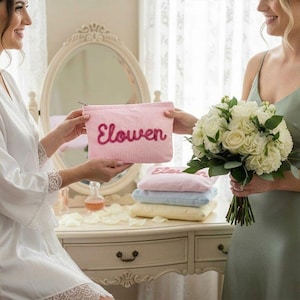 May include: A pink and white striped cosmetic bag with the name "Elowen" embroidered in a cursive font. The bag is being held by a person wearing a white robe. A bouquet of white roses is held by another person wearing a green dress.