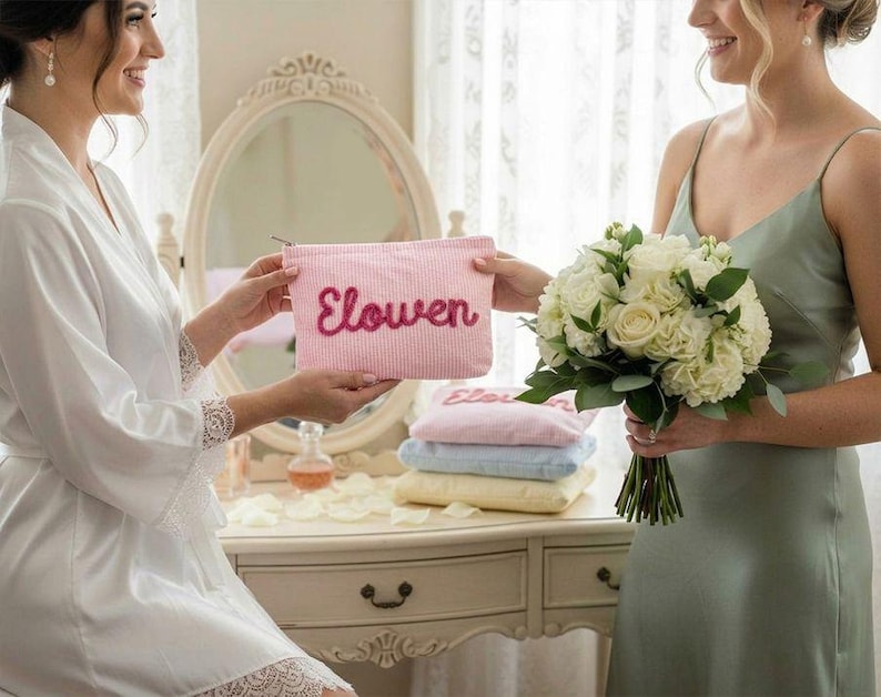 May include: A pink and white striped cosmetic bag with the name "Elowen" embroidered in a cursive font. The bag is being held by a person wearing a white robe. A bouquet of white roses is held by another person wearing a green dress.