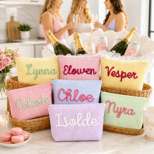 May include: A collection of colorful corduroy cosmetic bags, each embroidered with a different name in a cursive font. The bags are in shades of yellow, pink, blue, and green, and are arranged in a woven basket. Champagne bottles and glasses are in the background.
