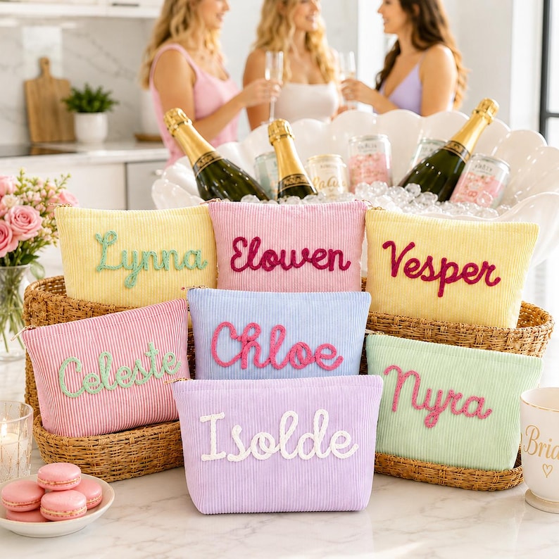 May include: A collection of colorful corduroy cosmetic bags, each embroidered with a different name in a cursive font. The bags are in shades of yellow, pink, blue, and green, and are arranged in a woven basket. Champagne bottles and glasses are in the background.