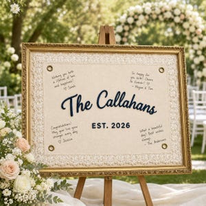 May include: A rectangular wedding guest book sign in a gold frame with a cream-colored fabric center featuring white lace trim. The text reads The Callahans EST. 2026 with handwritten messages in the corners and four brass grommets at the corners.