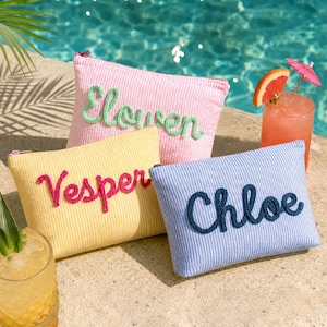 May include: Three corduroy cosmetic bags in pink, yellow, and blue, each with a name embroidered on the front: "Elowen", "Vesper", and "Chloe". The bags are rectangular with a zipper closure. A cocktail with a grapefruit slice and a small umbrella is in the background.