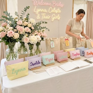 May include: A display of personalized gifts for a bridal party. The table features pastel-colored cosmetic bags with names embroidered on them, alongside tote bags, flowers, and a neon sign that reads "Welcome Brides: Lynna, Celeste & Their Girls."