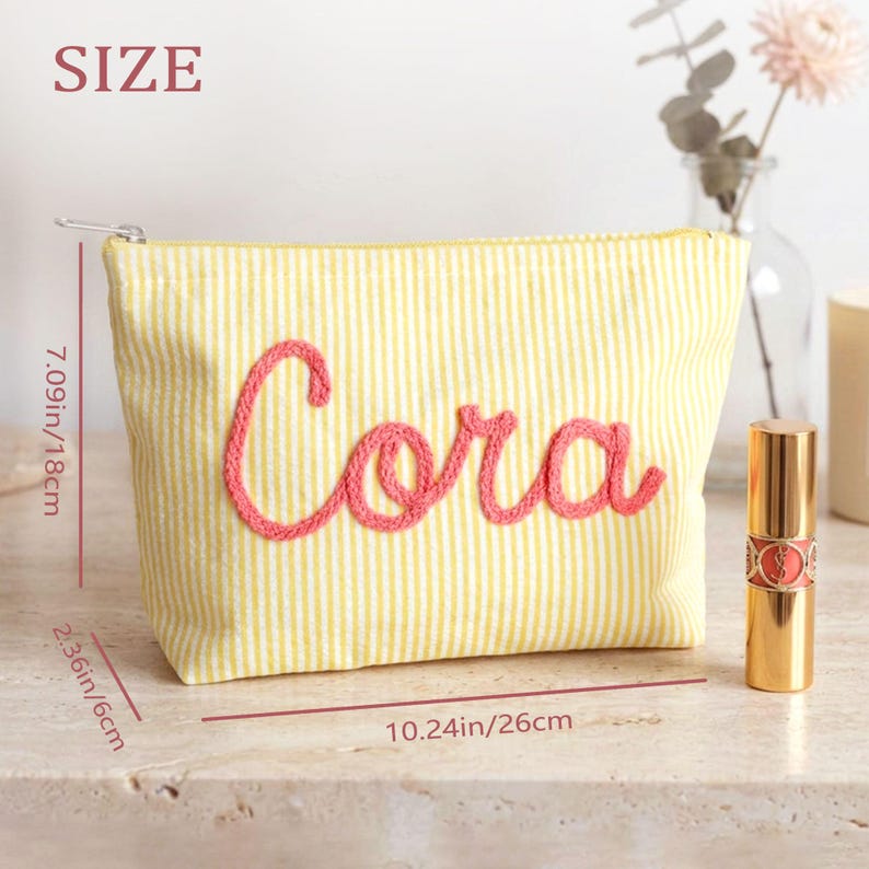 May include: Yellow and white striped cosmetic bag with the name "Cora" embroidered in pink. The bag is 7.09 inches (18cm) high, 10.24 inches (26cm) wide, and 2.36 inches (6cm) deep. A gold lipstick tube is next to the bag.