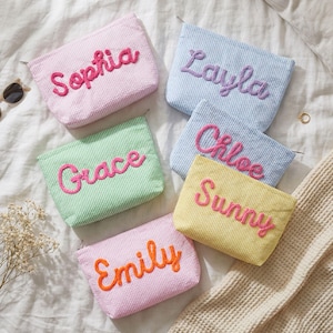 May include: A collection of personalized cosmetic bags in various pastel colors, including pink, blue, green, and yellow. Each bag features a different name embroidered in a contrasting color. The bags are arranged on a white textured surface.