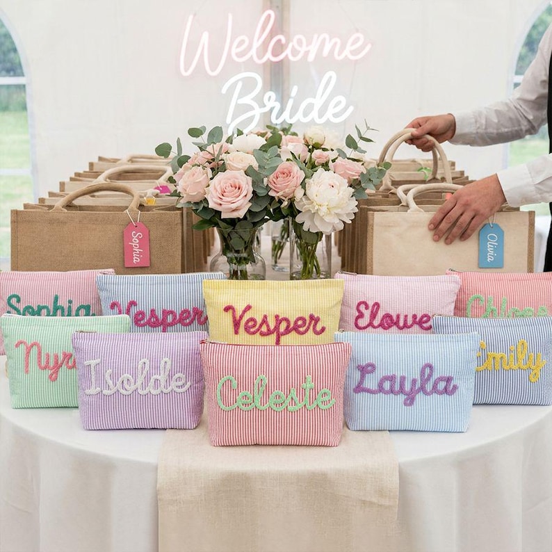 May include: A display of personalized cosmetic bags and tote bags, with the words "Welcome Bride" in neon. The cosmetic bags are striped in various pastel colors, each embroidered with a name. Tote bags are in the background.
