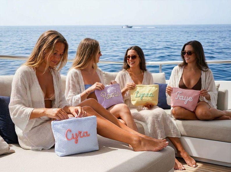 May include: Four women on a boat hold personalized cosmetic bags. The bags are pastel colors: light blue, lavender, yellow, and pink. Each bag has a name embroidered in a contrasting color. The background shows the ocean and a clear sky.