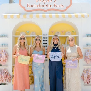 May include: Four women stand in front of a white and yellow outdoor display with the text "Vesper's Bachelorette Party." Each woman holds a personalized pouch. The pouches are pastel colors with names embroidered on them.