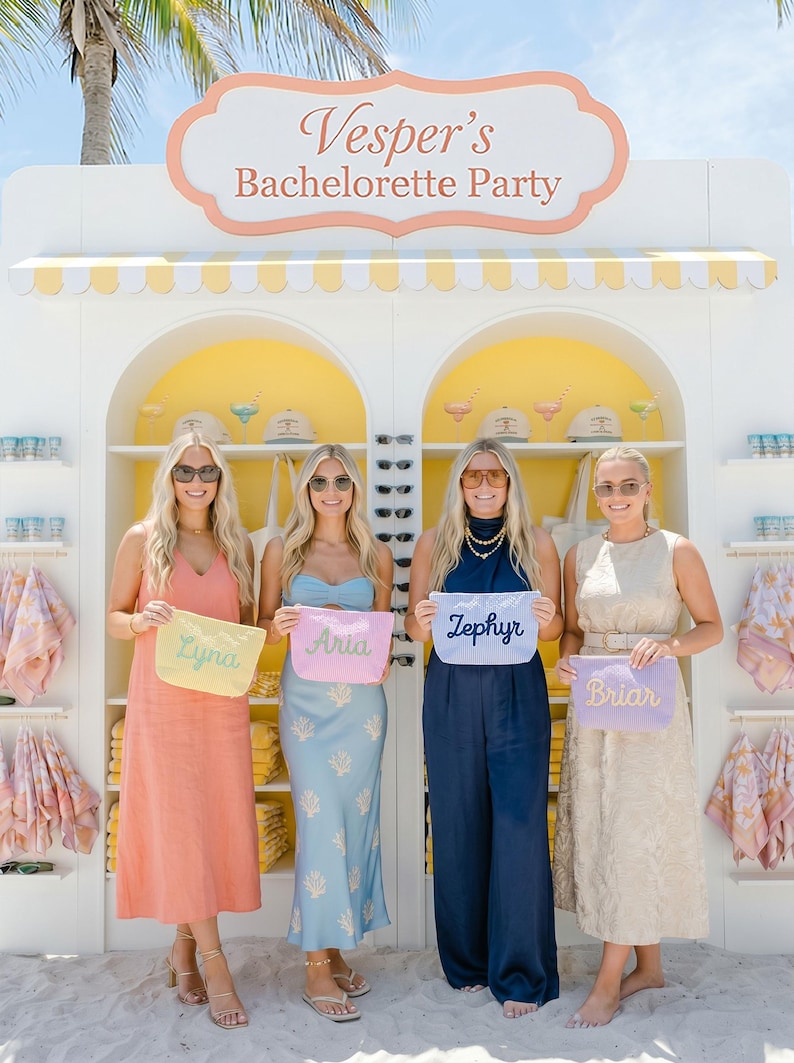 May include: Four women stand in front of a white and yellow outdoor display with the text "Vesper's Bachelorette Party." Each woman holds a personalized pouch. The pouches are pastel colors with names embroidered on them.
