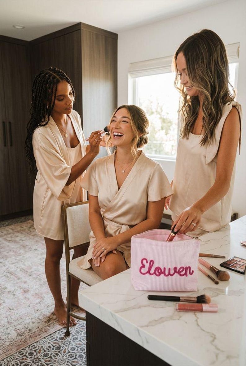 May include: Three people in matching beige robes, one applying makeup to another. A pink striped makeup bag with the name "Elowen" embroidered on it sits on a white countertop. Makeup brushes and products are visible.