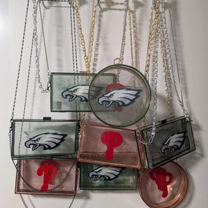 Może przedstawiać: Kolekcja przezroczystych akrylowych torebek z łańcuszkami. Niektóre torby mają logo Philadelphia Eagles, inne czerwone logo "P". Torby mają różne kształty, w tym prostokątne i okrągłe.