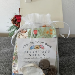May include: A clear, drawstring bag containing decoupage shells and fabric. The bag includes a label that reads "Create Your Own Decoupage Shells." The bag is decorated with a small pinecone and a red and black plaid ornament.