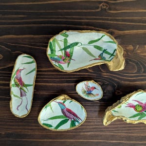 Puede incluir: Colección de platos decorativos de concha con borde dorado. Cada plato presenta un diseño único de colibrí con hojas de bambú verdes. Los colibríes son en tonos de rojo, rosa, azul y verde.