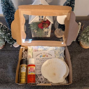Könnte beinhalten: Eine Bastelbox mit einem transparenten Fenster, die eine weiße Muschel, Farbtuben und eine Flasche Mod Podge enthält. Die Box enthält auch ein kleines dekoratives Haus und Grünzeug. Der Text "Create Your Own Decoupage Shell" ist sichtbar.