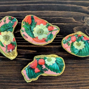 May include: Four decorative oyster shells with a strawberry and floral design. The shells feature a pink background with red strawberries, green leaves, and white flowers. The edges are trimmed with gold paint.
