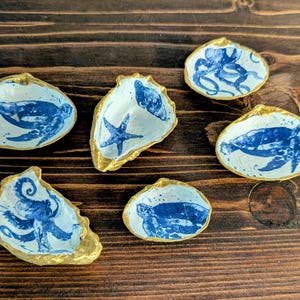 Peut inclure: Ensemble de six petits plats en forme de coquillage, bordés d'or. Chaque plat présente un motif marin bleu et blanc, avec des tortues, des pieuvres et des étoiles de mer. Posés sur une surface en bois sombre.