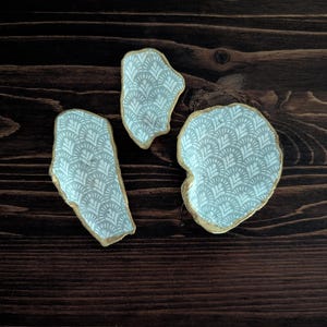Può includere: Tre piatti decorativi in ceramica con bordi irregolari. Ogni piatto presenta un interno a fantasia blu chiaro e bianco e un bordo dorato. I piatti sono disposti su una superficie di legno marrone scuro.