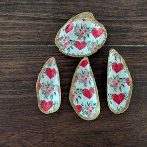 May include: Four seashell trinket dishes. The shells are painted white with red hearts and pink floral designs. The edges are trimmed with gold paint. The shells are arranged on a dark brown wooden surface.