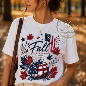 Op de afbeelding: Een wit t-shirt met korte mouwen en een grafische print van een pompoen met het patroon van de Amerikaanse vlag, omringd door rode en blauwe herfstbladeren, sterren en de tekst Fall SEASON EST. 2026 met een gestileerd Amerikaans vlagontwerp.