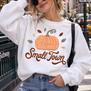 Op de afbeelding: Een wit sweatshirt met lange mouwen en een afbeelding van een lichtoranje pompoen met een groene steel, omringd door kleine bruine en groene blaadjes. Onder de pompoen staat de tekst Small Town in een donkerbruin cursief lettertype.