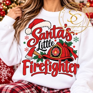 Op de afbeelding: Een wit sweatshirt met lange mouwen en een grafische print met de tekst Santa's Little Firefighter. Het ontwerp bevat een rode kerstmuts, een rode brandweerhelm met een vlamembleem, een bijl, groene hulstbladeren en rode sneeuwvlokken.