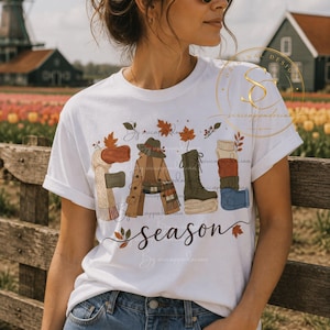Op de afbeelding: Een wit t-shirt met korte mouwen en een grafische print die het woord FALL vormt met diverse kledingstukken zoals een sjaal, een jas en laarzen, met het woord season in cursief eronder en verspreide herfstbladeren.