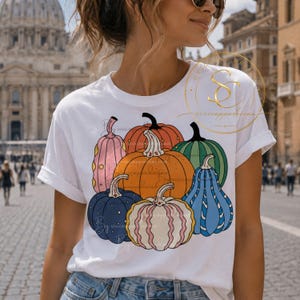 Op de afbeelding: Een wit T-shirt met korte mouwen en een grafische print van zeven gestileerde pompoenen in diverse kleuren, waaronder oranje, roze, groen en blauw. De pompoenen hebben verschillende patronen zoals strepen en stippen en zijn gegroepeerd.