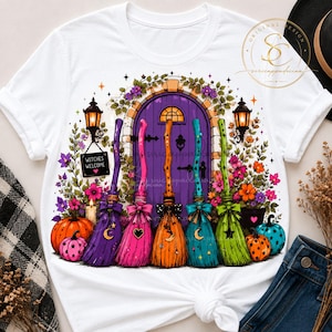 Op de afbeelding: Een wit t-shirt met een afbeelding van een paarse boogdeur omringd door bloemen, twee lantaarns en vijf kleurrijke bezems. Een klein zwart bordje hangt aan een lantaarn met de tekst Witches Welcome. Pompoenen staan aan de onderkant.